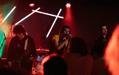 Three artists on stage with microphones and musical instruments performing to a crowd.